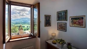 Una stanza con un'ampia finestra aperta che si affaccia su un panorama con montagne e cielo sereno. Alle pareti ci sono quadri appesi e un tavolino con una lampada e dei fiori.