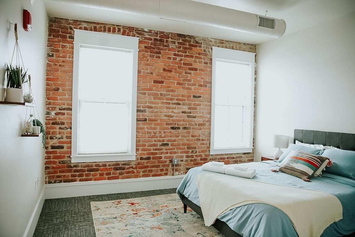 A bedroom with a brick wall, two windows, a bed with pillows, a bedside table, and a small plant on the wall.