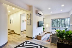 Uma sala de estar moderna com escada, TV de tela plana na parede e uma grande janela com vista para árvores.