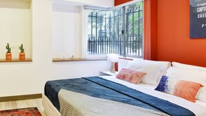 Ein Schlafzimmer mit einem Bett, einem Fenster mit Blick auf Bäume, einem Regal mit zwei Blumentöpfen und einem roten Teppich.