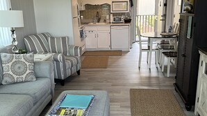 A modern living room with a grey sofa, a matching ottoman, and a dining area with a table and chairs. A kitchen with white cabinets and a microwave is visible in the background. - Atlantic Beach Condo within walking distance to the beach (Atlantic Beach)