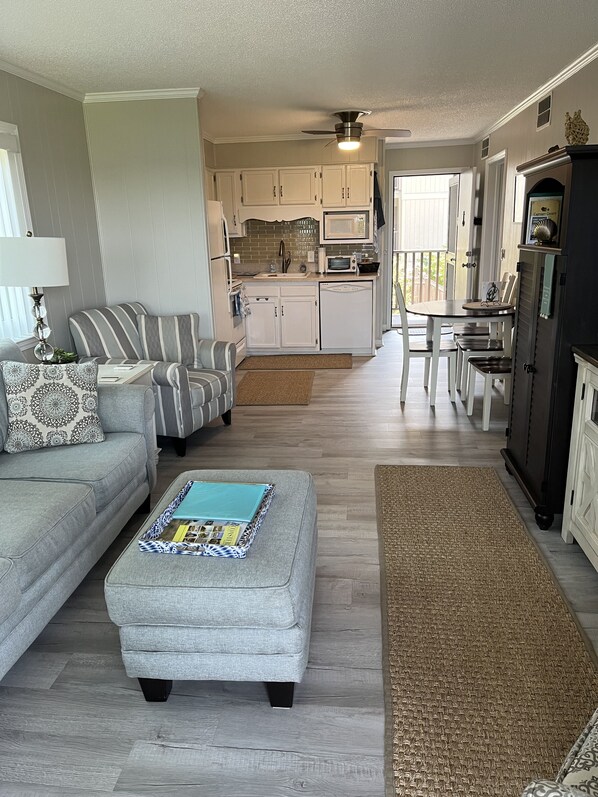 A modern living room with a grey sofa, a matching ottoman, and a dining area with a table and chairs. A kitchen with white cabinets and a microwave is visible in the background.