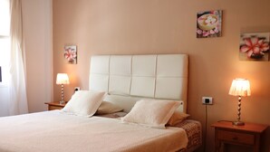 A neatly made bed with a headboard, two bedside tables with lamps, and floral wall art. - Casa Denise  (Playa San Juan)