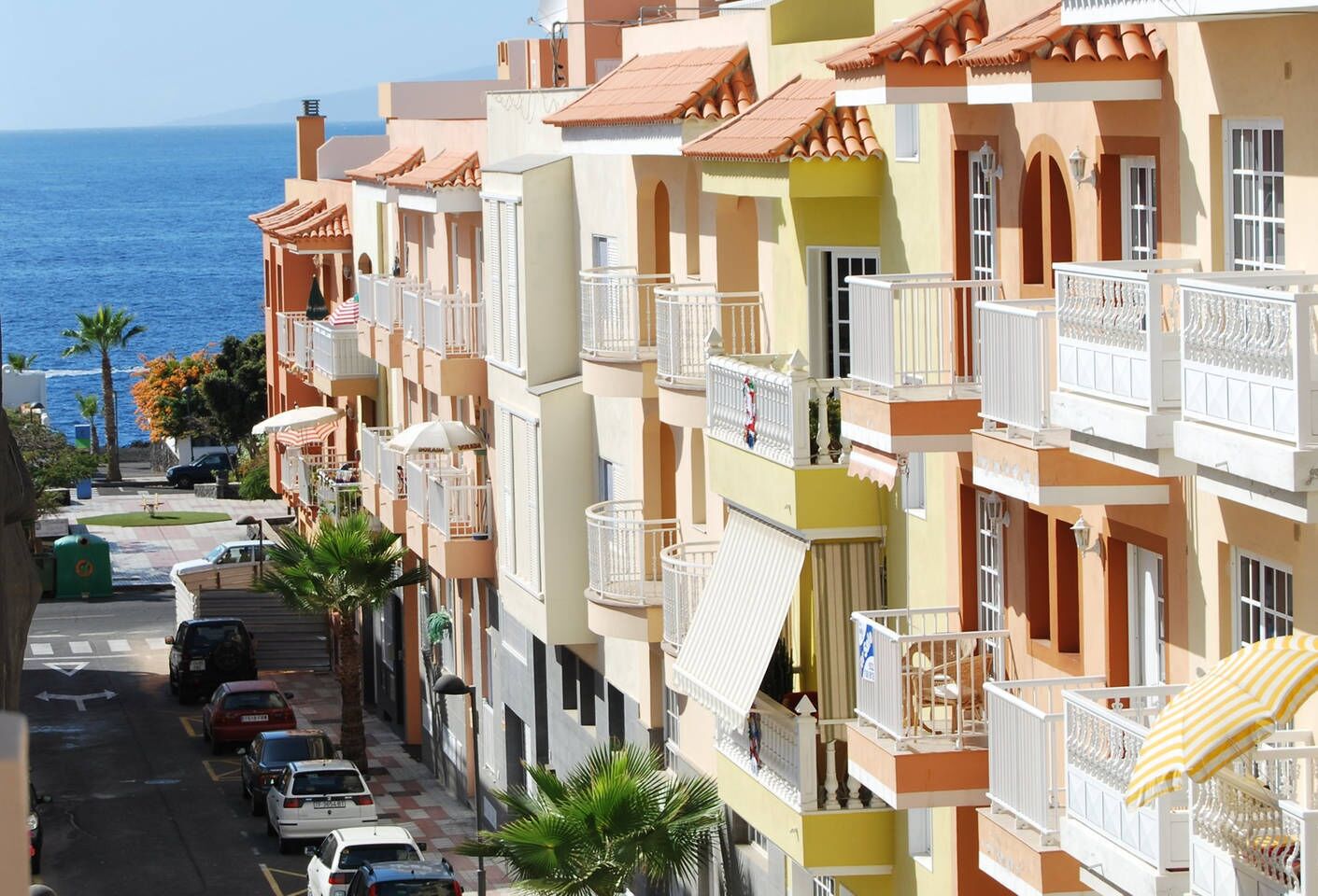 A row of colorful Mediterranean-style buildings with balconies, a street with parked cars, and a view of the sea.
