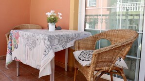 A dining area with a table covered by a patterned tablecloth, a wicker chair with a cushion, and a potted plant with white flowers. - Casa Denise  (Playa San Juan)