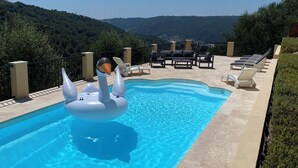 A swimming pool with two swan-shaped inflatable floats, surrounded by lounge chairs and a view of a hilly landscape. - Villa Avec Piscine Chauffée Nice Collines (Nice)