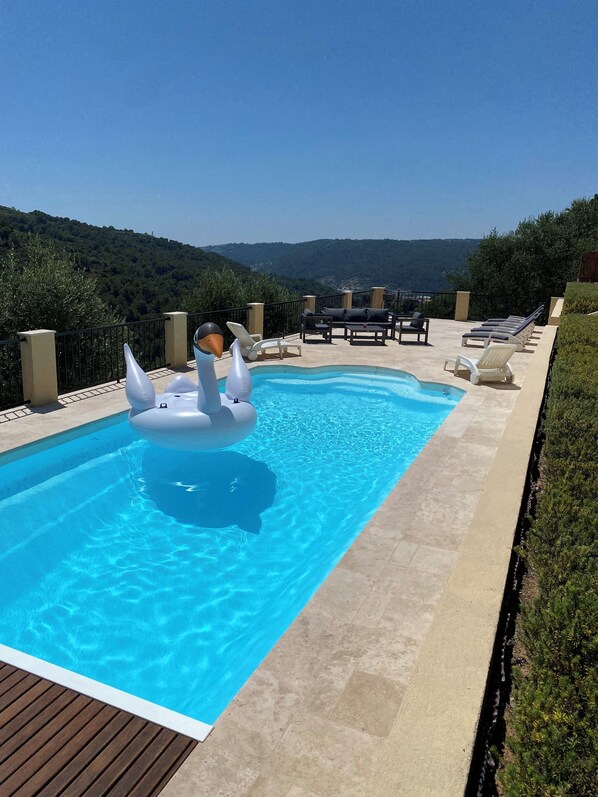 A swimming pool with two swan-shaped inflatable floats, surrounded by lounge chairs and a view of a hilly landscape. - Villa Avec Piscine Chauffée Nice Collines (Nice)