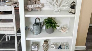 Un estante de madera blanca con objetos decorativos, incluyendo un cartel de bienvenida, una planta en maceta y una regadera de metal.
