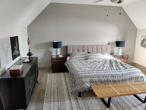 A bedroom with a bed, two bedside tables, a wooden bench, a dresser, a patterned rug, and a ceiling fan.