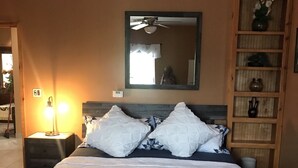 Un dormitorio con una cama grande, una mesita de noche con lámpara, un espejo sobre la cama y una estantería de madera con objetos decorativos.