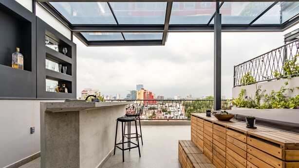 A modern rooftop bar with a glass enclosure, bar stools, and a wooden seating area.