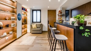 A modern hotel room with a wooden accent wall, a sofa, a kitchen area with bar stools, and a large window.