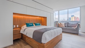 A modern bedroom with a large bed, a wooden headboard, and a city view through large windows.