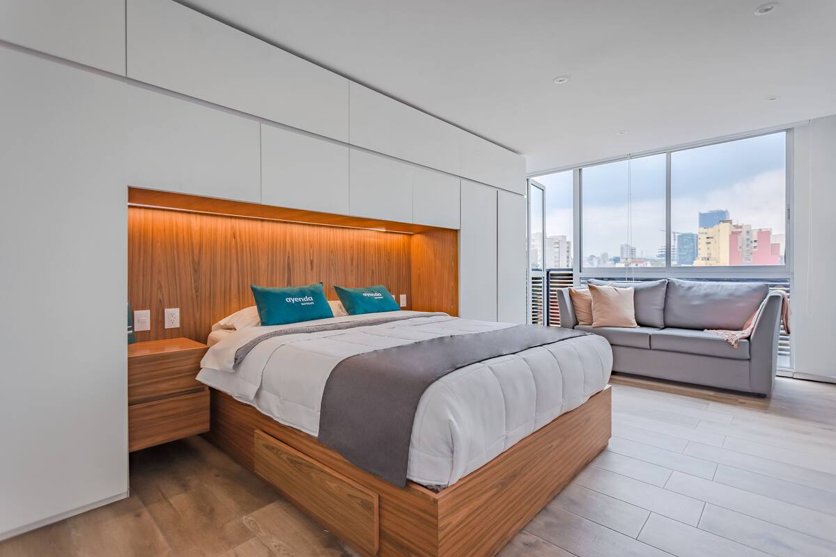 Een moderne slaapkamer met een groot bed, een houten hoofdbord en uitzicht op de stad door grote ramen.