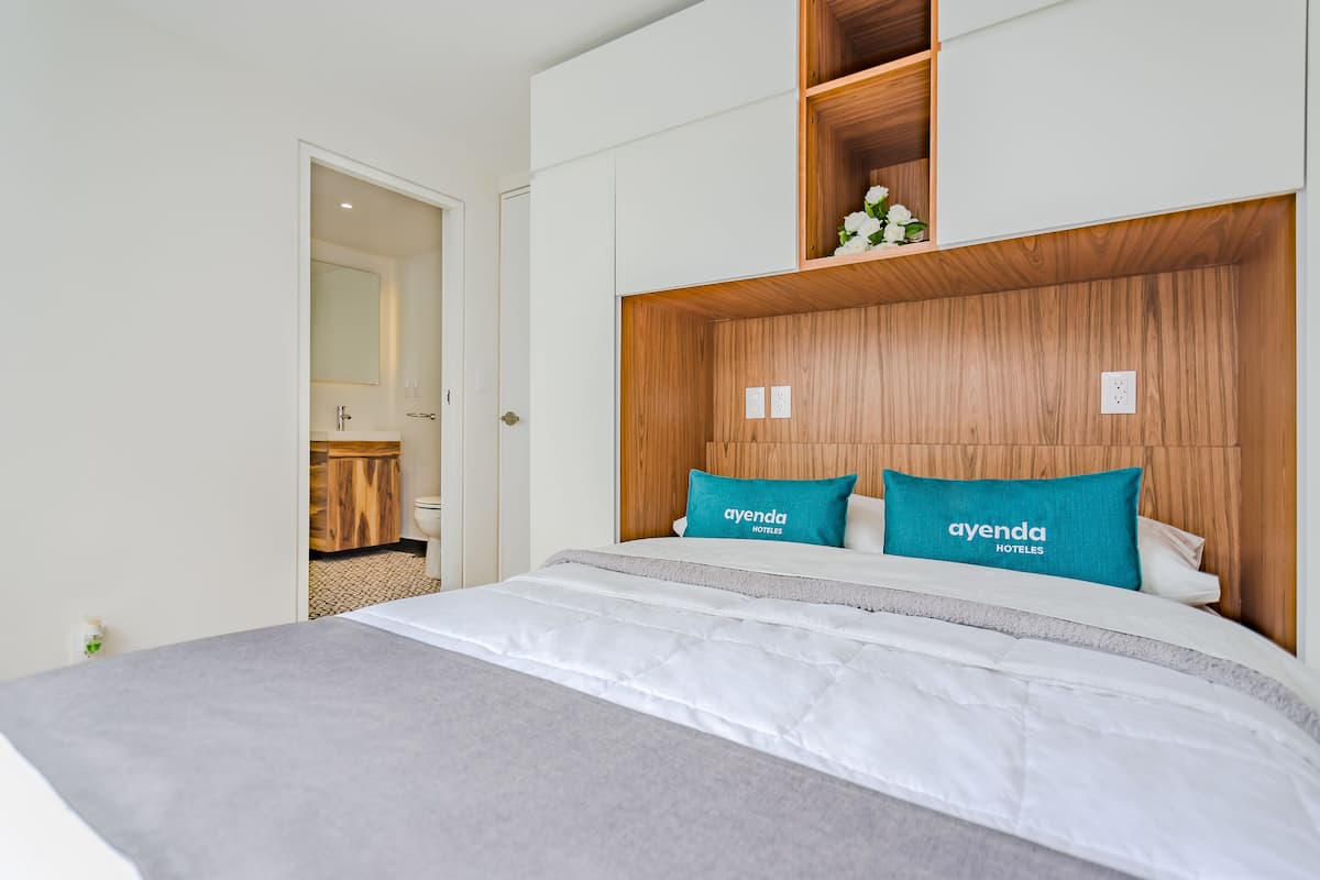 Een hotelkamer met een houten hoofdeinde, een bed met witte beddengoed en een teal kussen met de tekst 'ayenda HOTELES'.