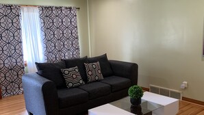 A living room with a grey sofa, patterned cushions, a white side table, and a small potted plant.
