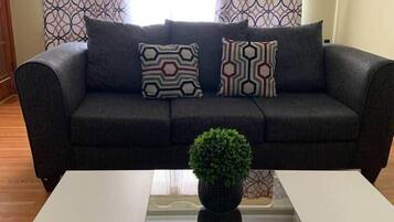 Una sala de estar con un sofá negro, cojines estampados, una mesa de centro de vidrio con una planta y cortinas con diseño.
