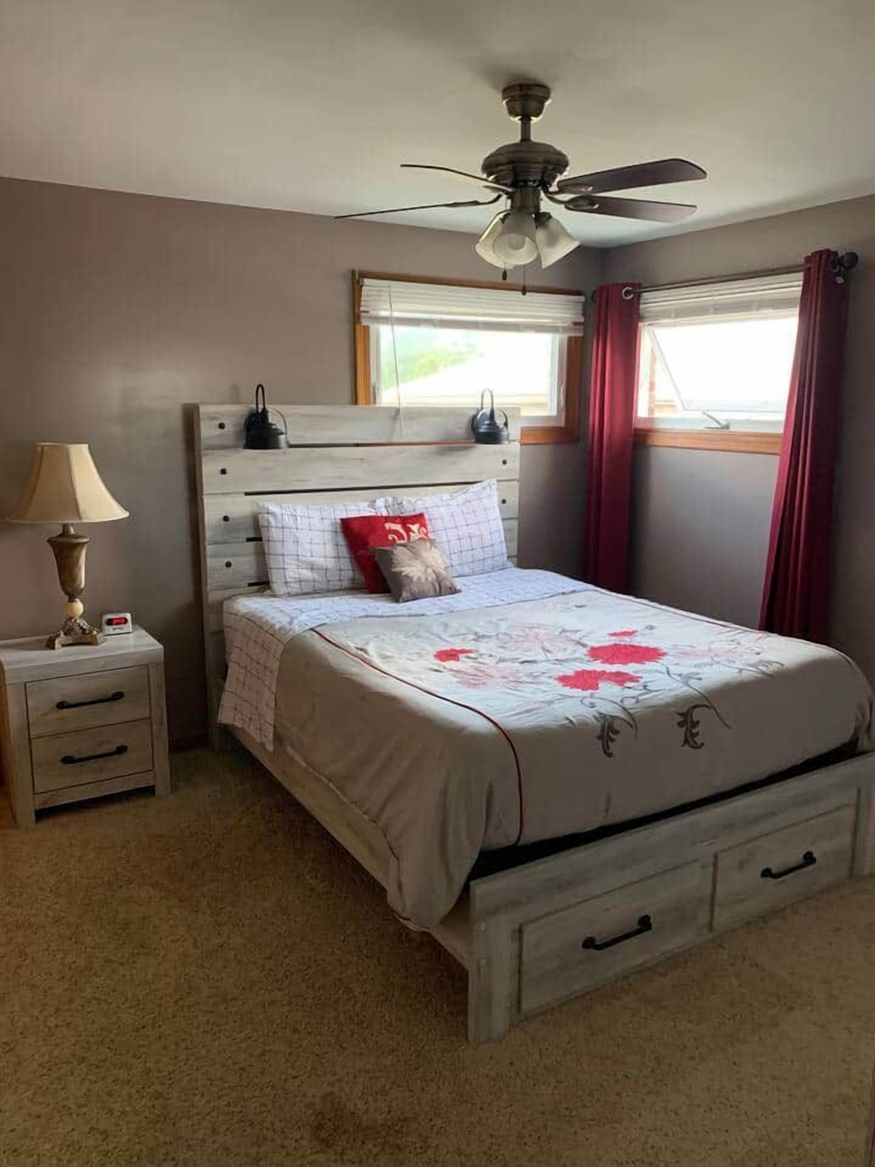 A bedroom with a bed, nightstand, lamp, ceiling fan, and window with curtains.