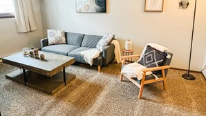 Un salon avec un canapé gris, une table basse en bois, un fauteuil en bois et un lampadaire. Deux œuvres d’art encadrées ornent le mur, et une fenêtre est agrémentée de rideaux.