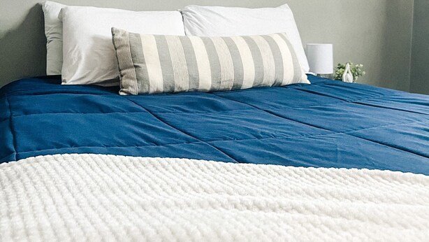 Une chambre avec une couvre-lit bleue, des oreillers blancs et une couverture blanche à texture. Deux reproductions encadrées de motifs botaniques ornent le mur.