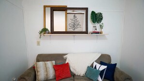 Una sala de estar con un sofá marrón, varios cojines, una obra de arte enmarcada y una planta sobre una estantería.