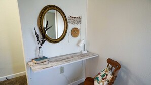 Un baño pequeño con tocador de madera, un espejo, un estante en la pared con un libro y un florero, y una silla con cojín estampado con flores.