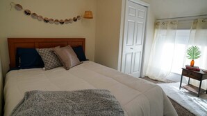 Un dormitorio con cama, almohadas, una colcha, una puerta, una ventana con cortinas, una pequeña mesa con una planta y adornos en la pared.
