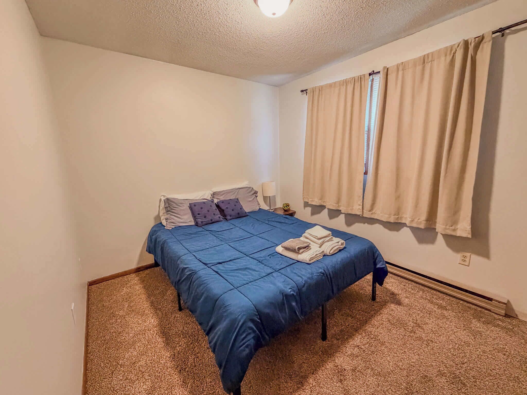 A bedroom with a bed, pillows, a nightstand, a lamp, and beige curtains.