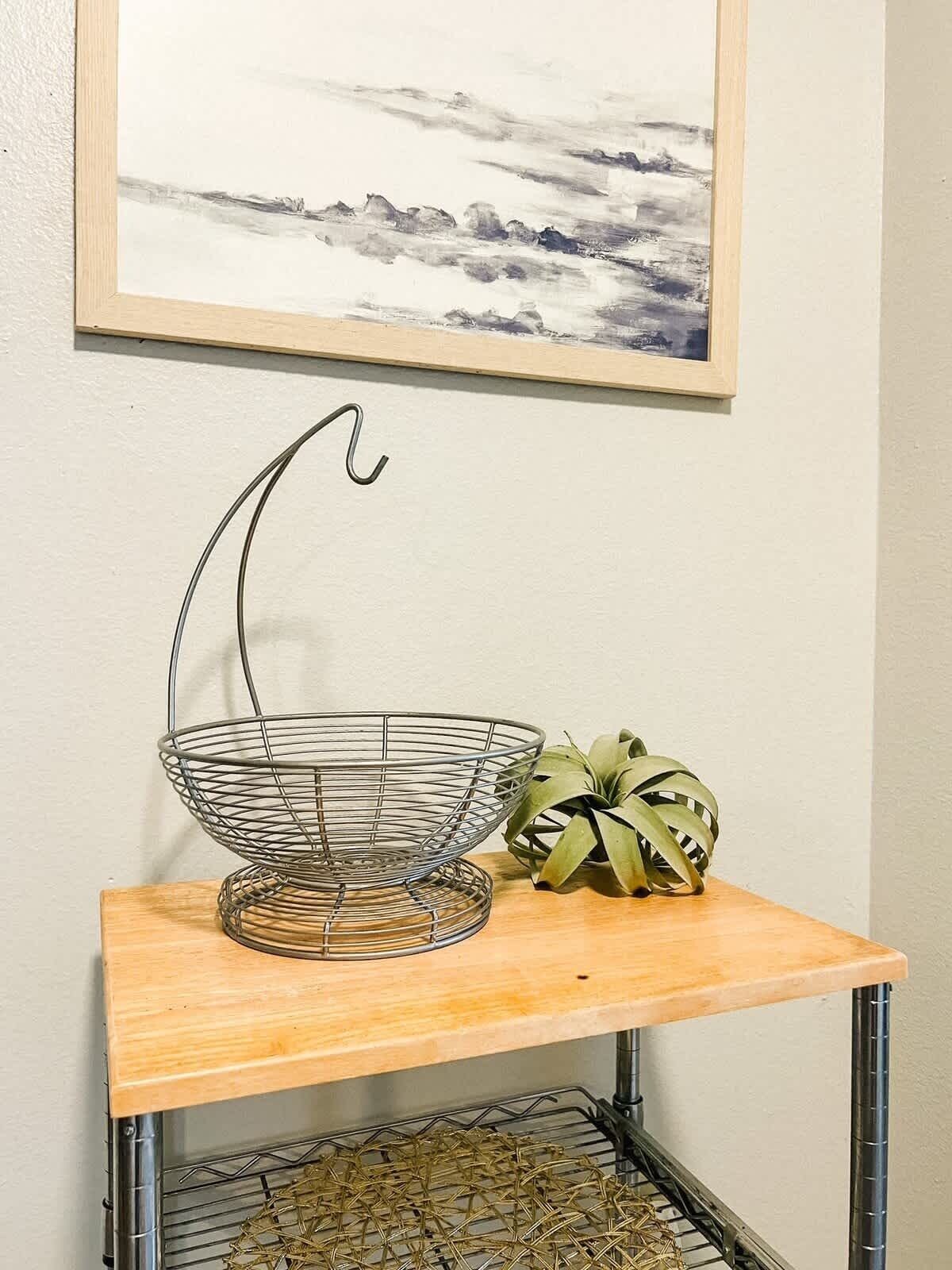 A metal wire basket on a wooden table with a plant beside it, under a framed artwork on the wall.