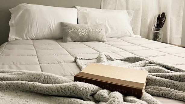 Ein ordentlich bezogenes Bett mit einer gesteppten Bettdecke, einer flauschigen weißen Decke und einem Kissen mit dem Aufdruck 'snuggle up'.