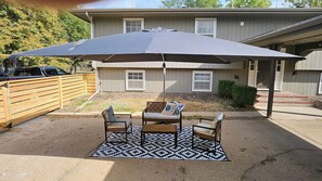 Une terrasse extérieure avec une table et des chaises, un grand parasol, et une maison en arrière-plan.
