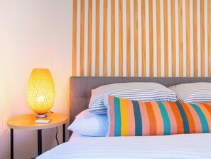 Una cama bien hecha con almohadas a rayas, una mesita de noche redonda con una lámpara y una pared con listones de madera verticales.