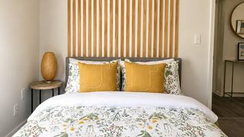 A bedroom with a bed, a headboard with vertical slats, a round mirror, and a small table with a lamp.
