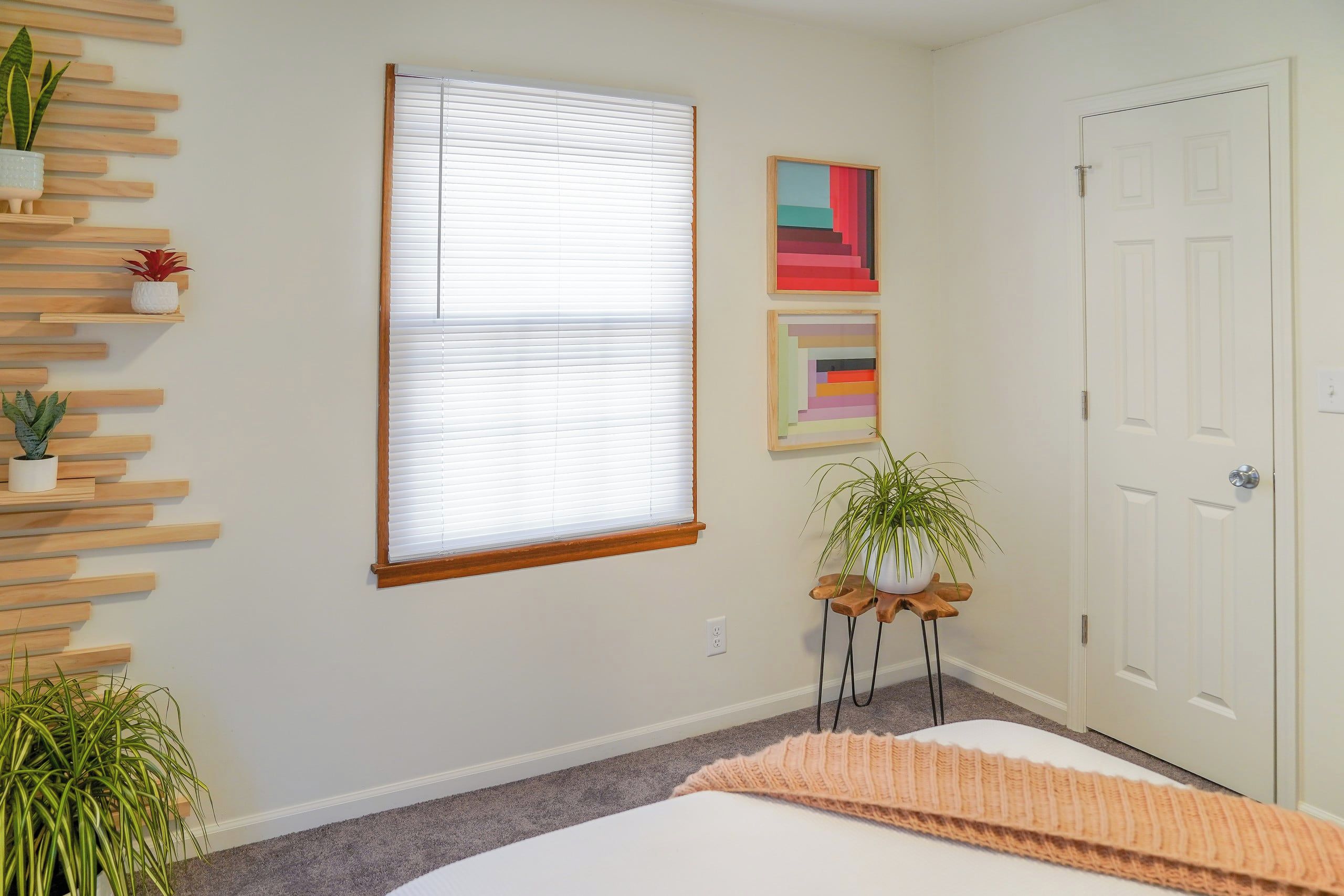 Une chambre à coucher avec un lit, une fenêtre avec des stores, une porte et une étagère fixée au mur, ornée de plantes.