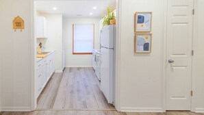 A narrow hallway leading to a kitchen with white cabinets, a refrigerator, and a window with blinds. - [3] StayAwhile: The Cozy Retreat (Lawrence)