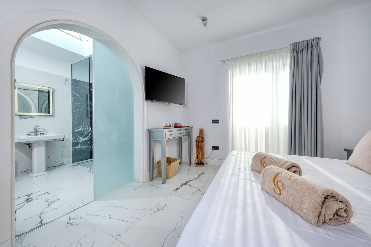 A modern hotel room with a large bed, a flat-screen TV, a vanity with a mirror, a sink, and a shower area.