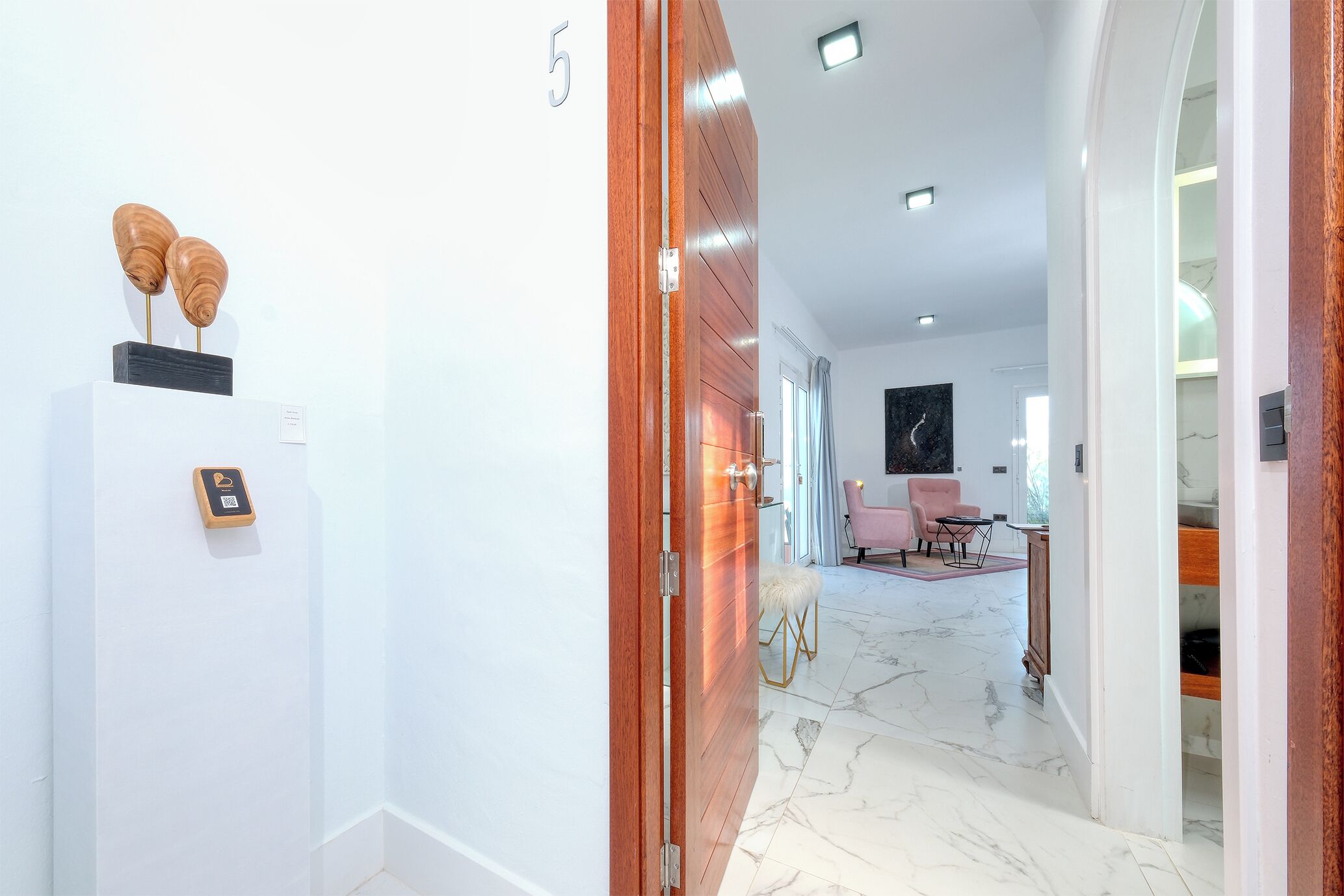 A modern hotel hallway with a marble floor, a wooden door, and a reception area with a pink chair and a small table.