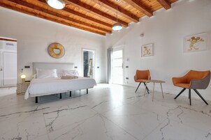 A spacious bedroom with a large bed, a wooden ceiling, and a marble floor. There are two chairs and a small table in the room.