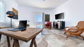 Una sala de estar moderna con una mesa de madera, una computadora portátil y una lámpara negra. Hay un televisor montado en la pared, una silla de madera y vistas al exterior a través de una puerta de cristal.