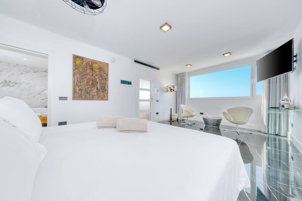 A modern hotel room with a large bed, a flat-screen TV, a large window, and a marble floor.