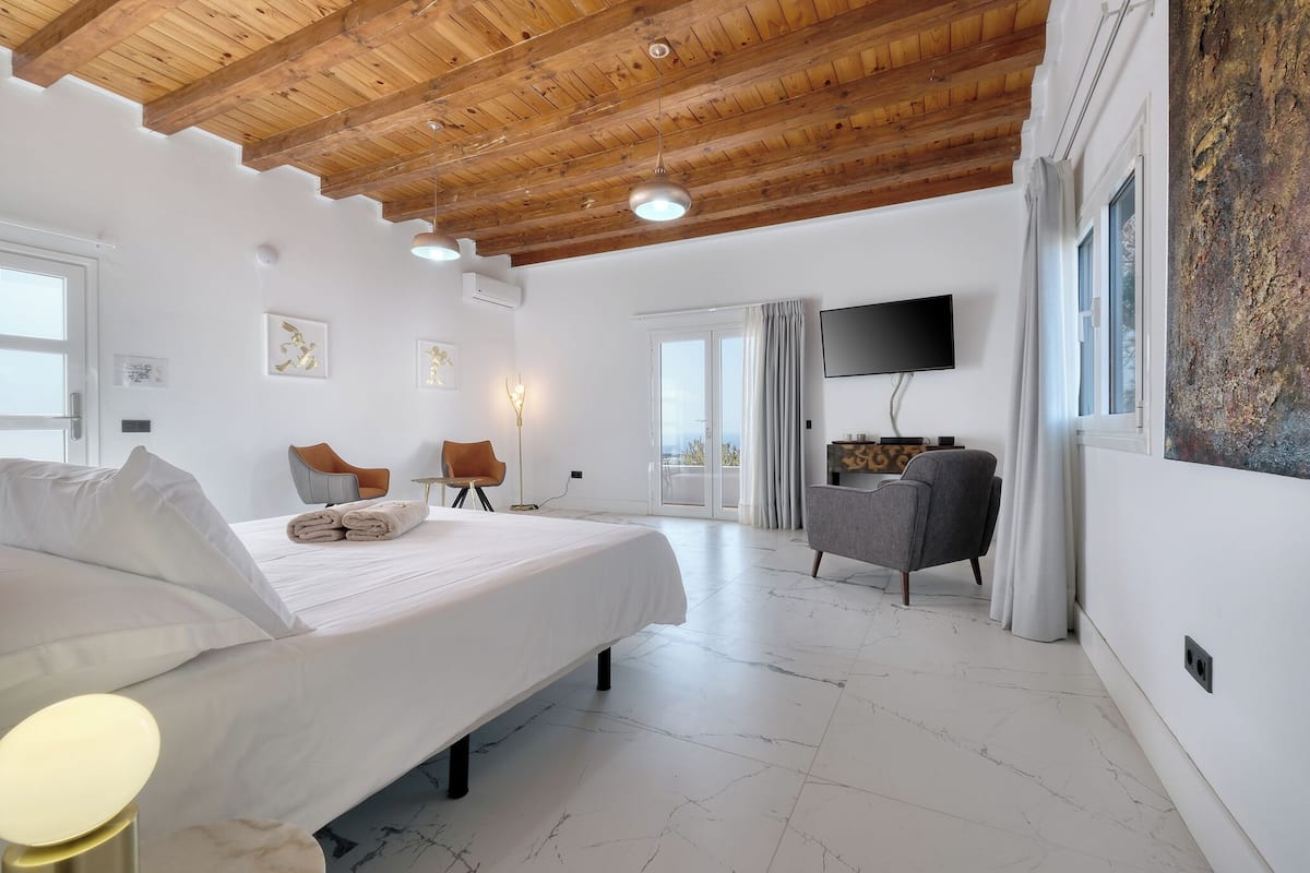 A modern hotel room with a large bed, a flat-screen TV, a seating area with chairs, and a balcony with a view.