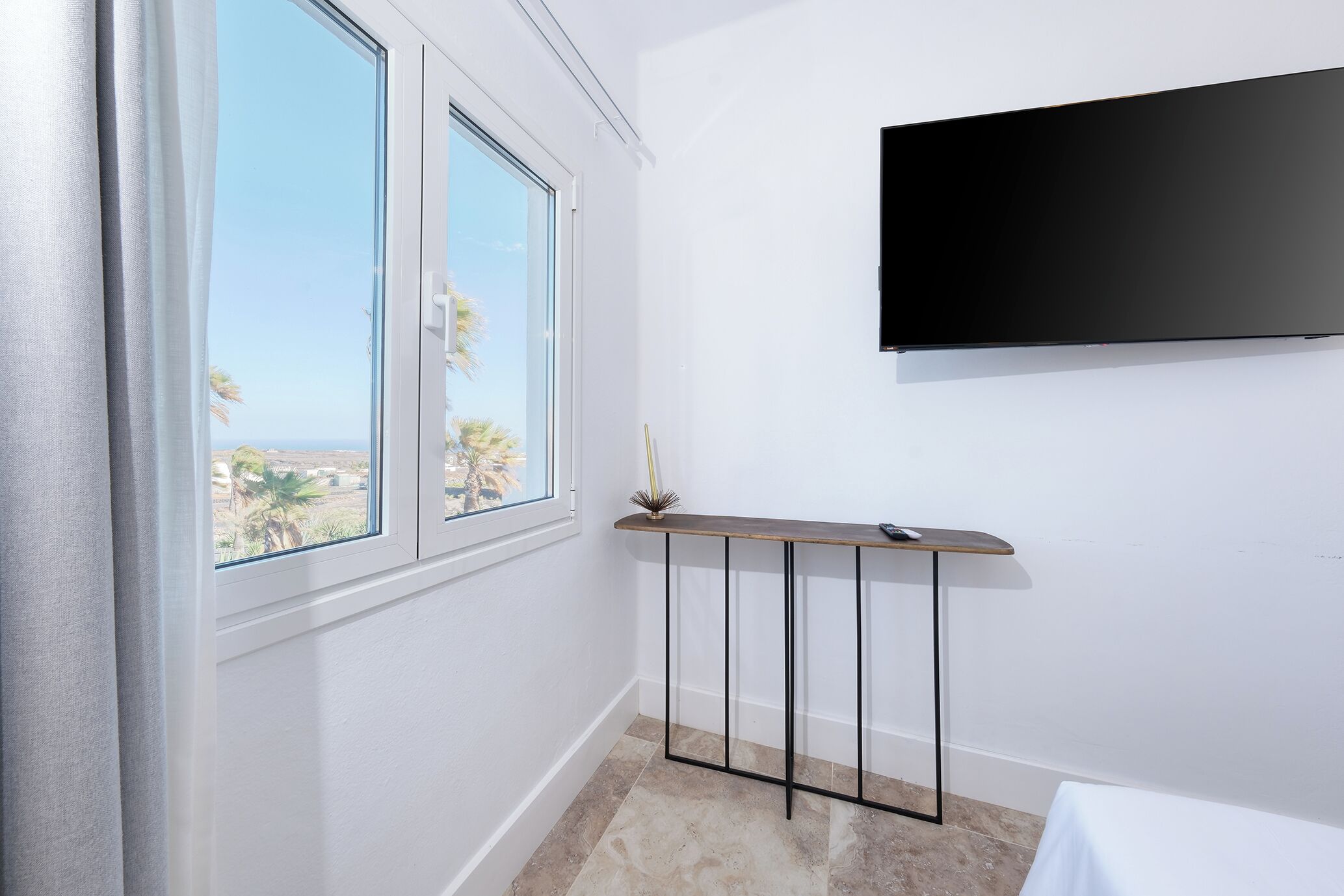 Una habitación con un ventanal grande, un televisor de pantalla plana en la pared y una mesita con un jarrón.