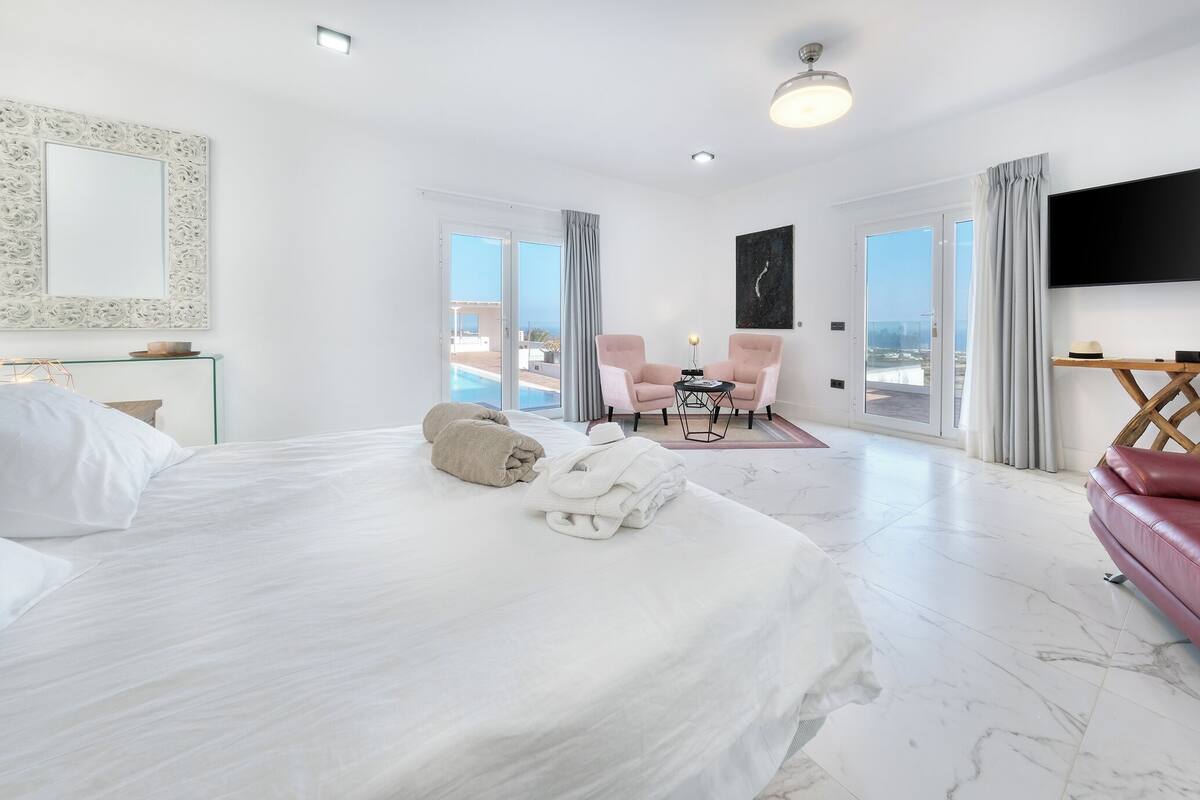 A hotel room with a large bed, two chairs, a TV, and a view of the ocean.
