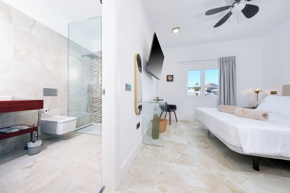 A modern hotel room with a glass shower, a bed with a white bedspread, a mounted TV, and a view of the outdoors.