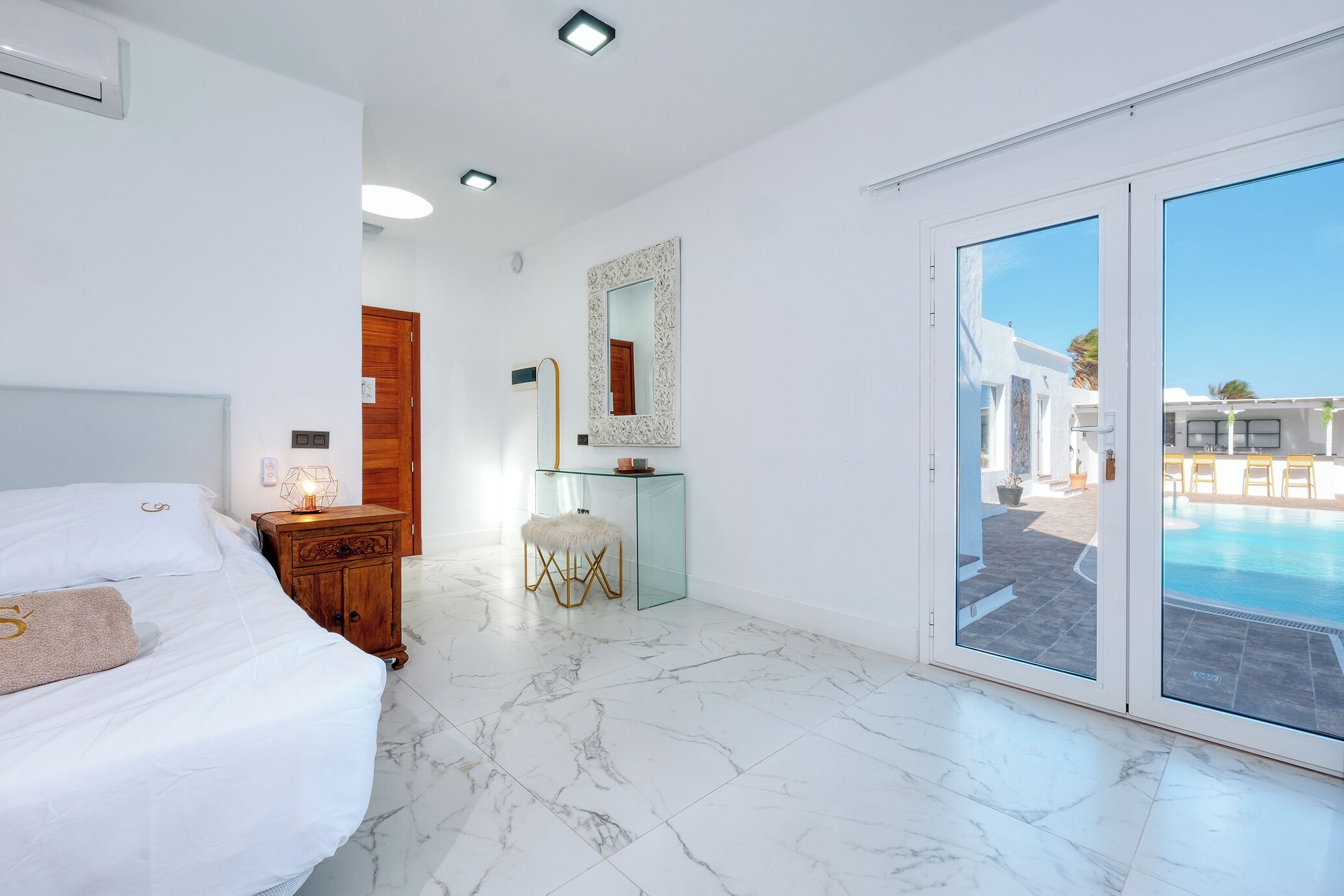 Quarto de hotel moderno com uma cama grande, uma mesinha de vidro, um espelho e vista para uma piscina.
