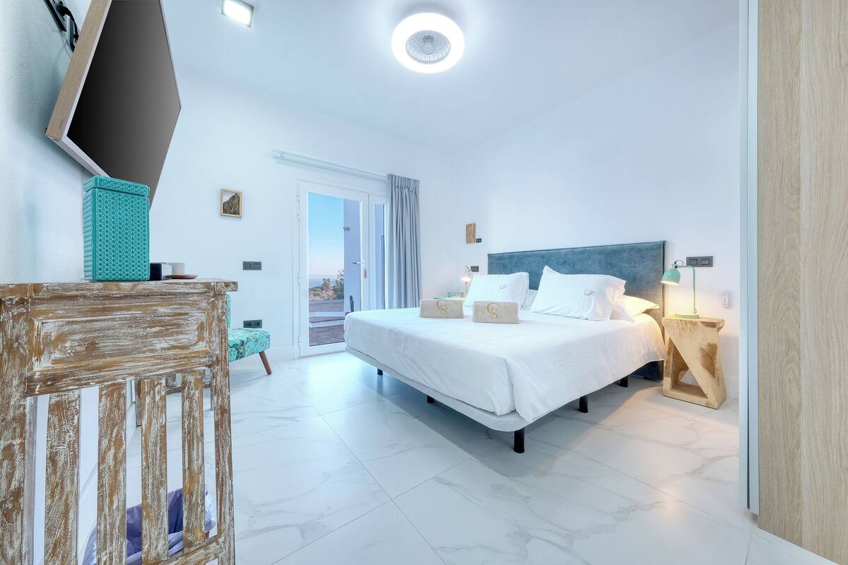 A modern hotel room with a large bed, a wooden headboard, a bedside table with a lamp, and a view of the outdoors through the door.