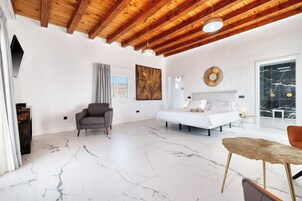 Una habitación amplia con un techo alto de madera, una cama blanca, un sillón gris, una mesita redonda pequeña y un cuadro abstracto grande en la pared.