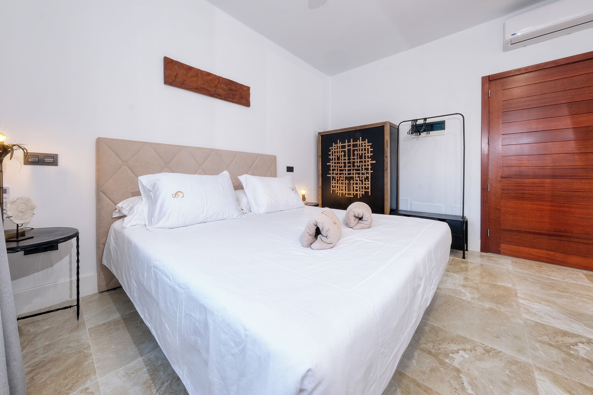 Una cama bien hecha con sábanas blancas y dos almohadones decorativos, un cabecero de madera, un equipo de aire acondicionado instalado en la pared y una puerta de madera.