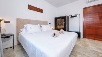 Una cama bien hecha con sábanas blancas y dos almohadones decorativos, un cabecero de madera, un equipo de aire acondicionado instalado en la pared y una puerta de madera.