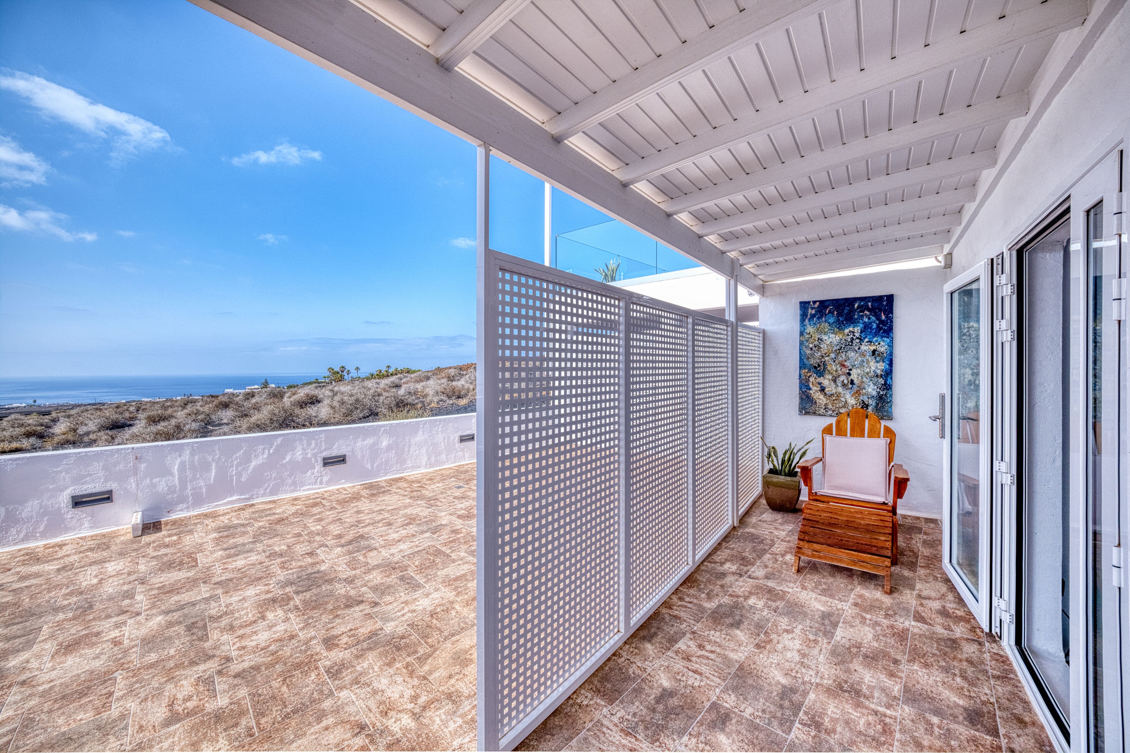 A covered balcony with a view of the ocean, a wooden chair, and a potted plant.